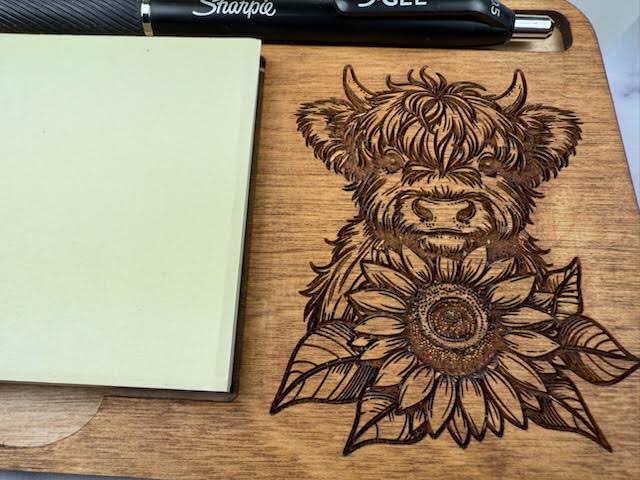 Wooden cutting board with a laser-engraved design of a cow and sunflower, next to a Sharpie pen. Sewing Bee & Laser Studio.