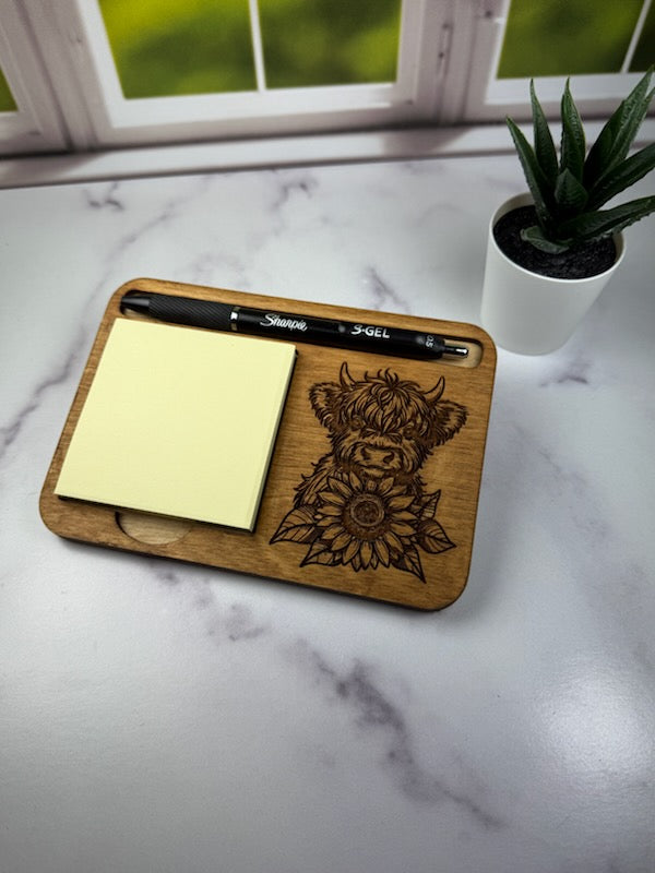 Wooden tray with laser-cut design, pen, and sticky notes on a marble surface with a plant in the background. Sewing Bee & Laser Studio.