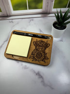 Wooden tray with laser-cut design, pen, and sticky notes on a marble surface with a plant in the background. Sewing Bee & Laser Studio.