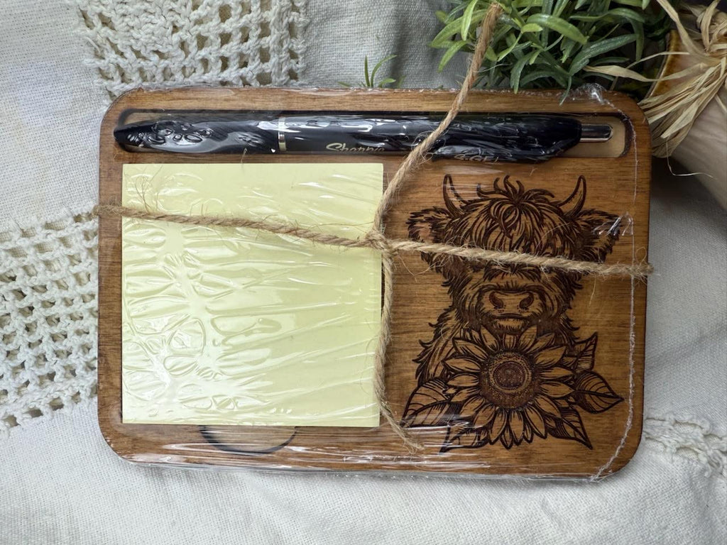 Wooden cutting board with laser-cut design of a highland cow and sunflower, tied with twine, on a textured surface. Shrink wrapped as mailed. Sewing Bee & Laser Studio.