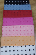 Set of 7 fabric fat-quarters with X-O patterns on various colors displayed on a wooden surface. Sold by Sewing Bee Studio.