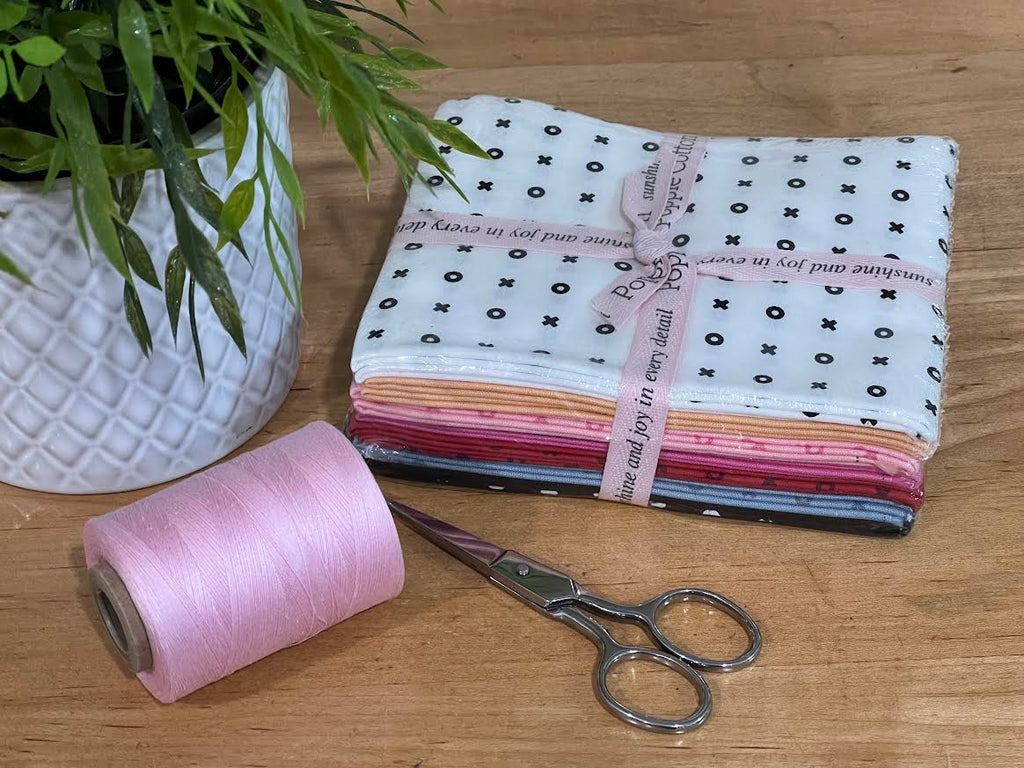 Fabric fat-quarter bundle with 7 fabric colors all with X-O patterns, with a spool of coordinating pink thread, and scissors on a wooden surface with a plant in the background. Bundle is sold by Sewing Bee Studio.