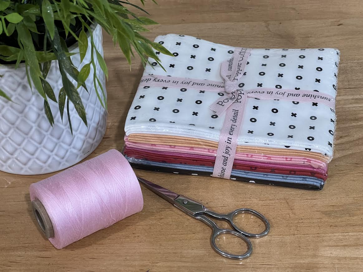 Fabric fat-quarter bundle with 7 fabric colors all with X-O patterns, with a spool of coordinating pink thread, and scissors on a wooden surface with a plant in the background. Bundle is sold by Sewing Bee Studio.