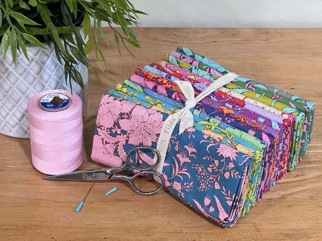 Bundle of colorful "Bloomsville" fabric fat-quarters on a table with complementary accents around it. Collection is sold by Sewing Bee Studio.