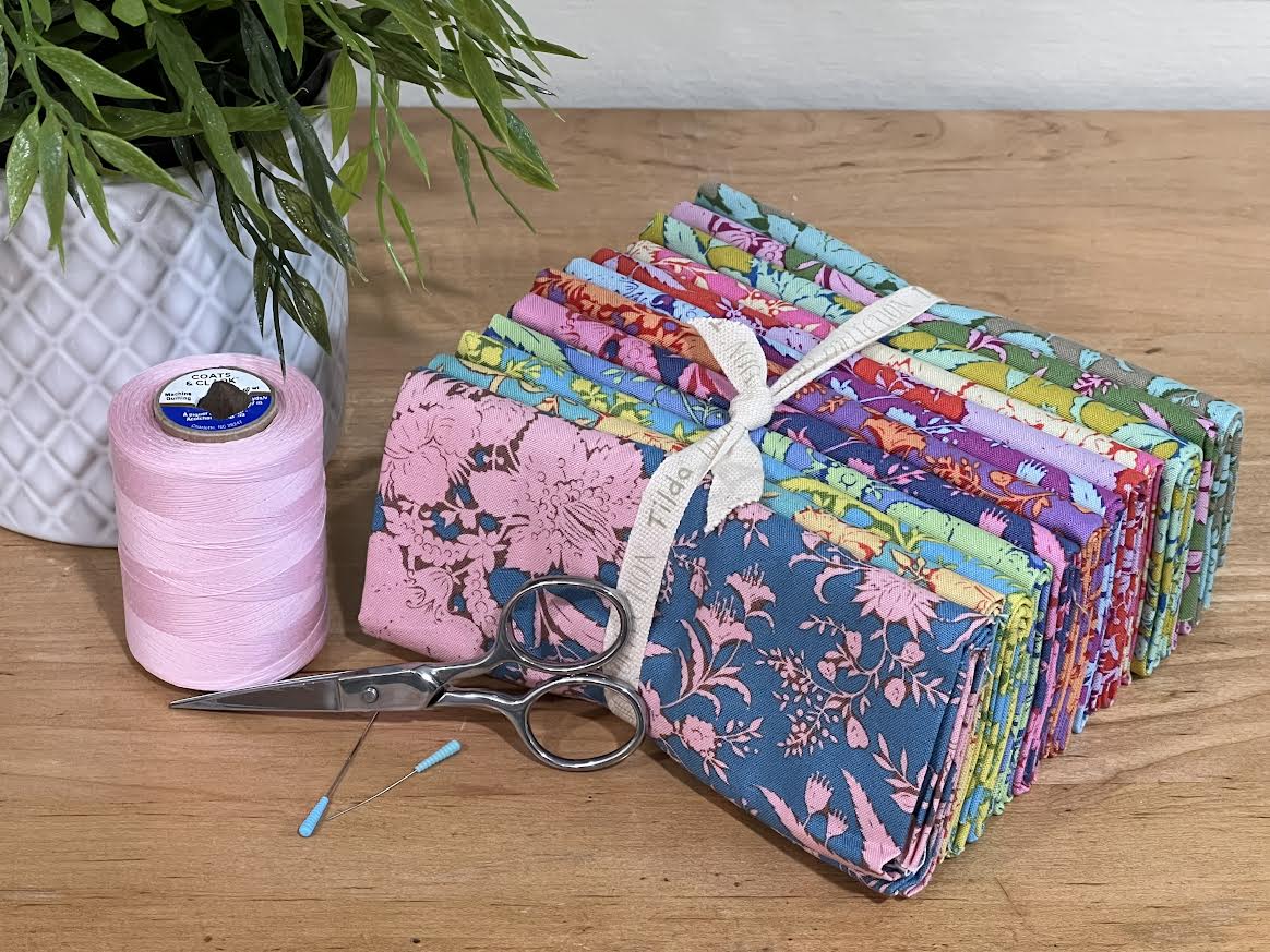 Bundle of colorful "Bloomsville" fabric fat-quarters on a table with complementary accents around it. Collection is sold by Sewing Bee Studio.