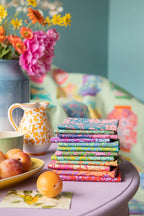 Stack of colorful fabric fat-quarters on a table with complementary accents in the background. Collection is sold by Sewing Bee Studio.